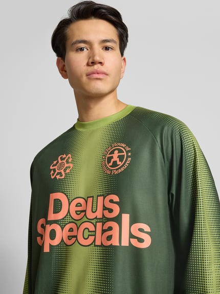 Männermodel trägt Trikot mit Label-Print und Rundhalsausschnitt von DEUS EX MACHINA in Regular-Fit, Longsleeve, grün-orange, Nahaufnahme Oberkörper. Rückenlänge bei Größe M: 71 cm, Ärmellänge bei Größe M: 65 cm.