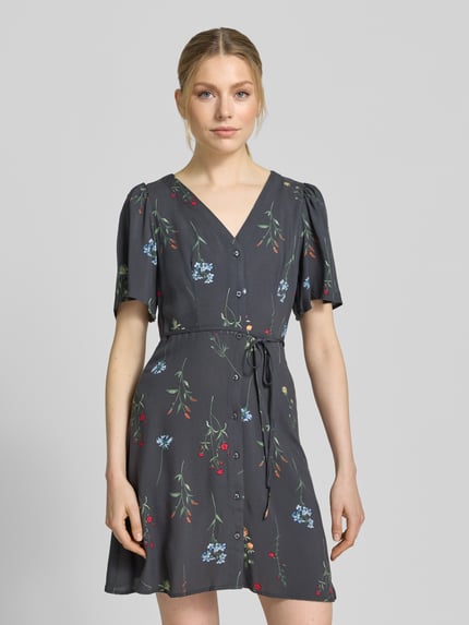 Vrouwenmodel draagt Leicht ausgestelltes Minikleid aus reiner Viskose Modell 'ALBA' van Vero Moda, donkergrijs met bloemendessin, uni-optiek, mini lengte, v-hals, tailleriempje, knoopsluiting, korte wijde mouwen, materiaal viscose, frontale zichtbare volledige lichaamsopname.