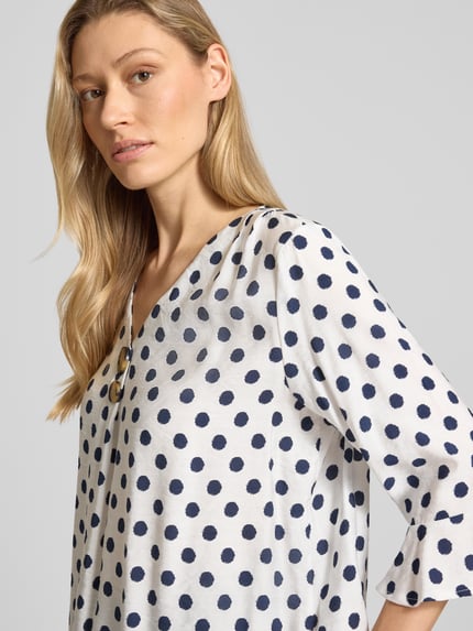 Modelka nosi Blusenshirt mit Viskose-Anteil von Apricot, mieszanka materiałów z dodatkiem wiskozy, prosty krój, dekolt w serek, ozdobne guziki, rękaw o dł. 3/4, długość tyłu w rozmiarze S: 67 cm, długość rękawów w rozmiarze S: 41 cm, biały materiał w granatowe grochy, faktura gładka; zbliżenie na górną część ciała, widok z przodu.