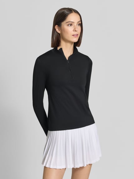 Frauenmodel trägt schwarzes Slim Fit Longsleeve mit Troyer-Kragen und Reißverschluss, dazu weißen Faltenrock, Modell 'FEATHER' von Fabletics, Rückansicht schräg von vorne, Rückenlänge bei Größe S: 54 cm.