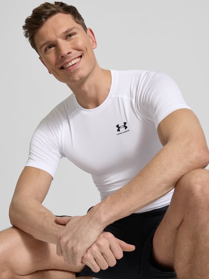 Męski model trägt Slim Fit T-Shirt mit Logo-Print von Under Armour, elastyczna mieszanka materiałów, krój slim fit, okrągły dekolt, długość tyłu w rozmiarze M: 62 cm, jasna biel, czarny nadruk z logo, gładka tekstura, nahaufnahme oberkörper.