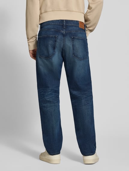 Männermodel trägt Tapered Fit Jeans aus Baumwoll-Mix von Tommy Jeans in Dunkelblau mit sichtbaren Nähten, Ganzkörperansicht Rückansicht. Herren Tapered Fit Jeans von Tommy Jeans.
