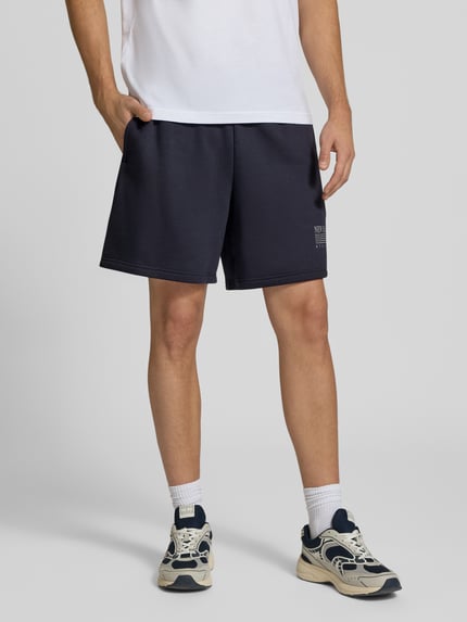 Männermodel trägt Sweatshorts aus Baumwoll-Mix von New Balance in Dunkelblau, gerader Schnitt, elastischer Bund, Label-Print, Nahaufnahme Unterkörper.