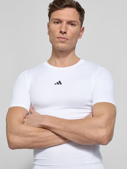 Męski model nosi T-Shirt mit Label-Print od adidas Training, biały kolor, materiał z domieszką stretchu, krój slim fit, całkowite ujęcie z przodu.