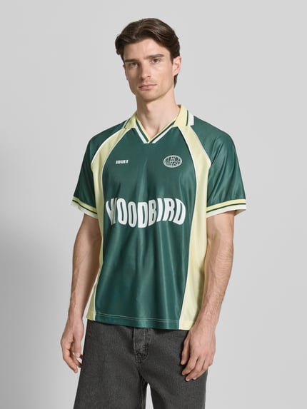 Männermodel trägt Poloshirt mit Label-Print Modell 'Taka' von WOODBIRD in Grün mit Polokragen, lockere Passform, Kurzärmelig, Ganzkörperansicht von vorne.