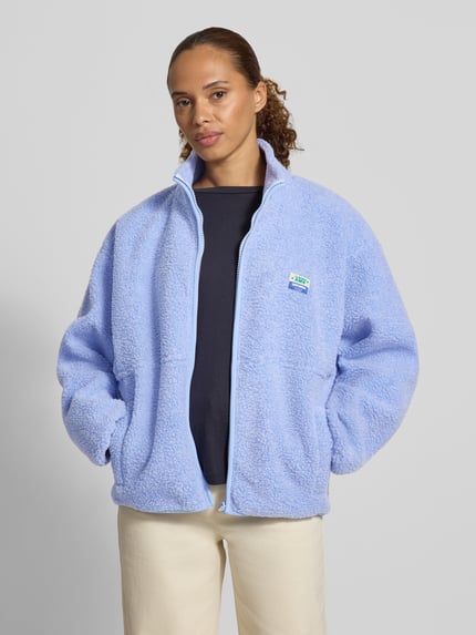 Frauenmodel trägt Regular Fit Jacke aus Teddyfell Modell 'Hoktown' von American Vintage in Hellblau mit Stehkragen und Logo-Patch, Ganzkörperansicht von vorne. Damen Jacke, Reißverschluss, Eingrifftaschen, XS/S: Rückenlänge 62 cm, Schulterbreite 54 cm, Brustweite 46 cm, Ärmellänge 55 cm.