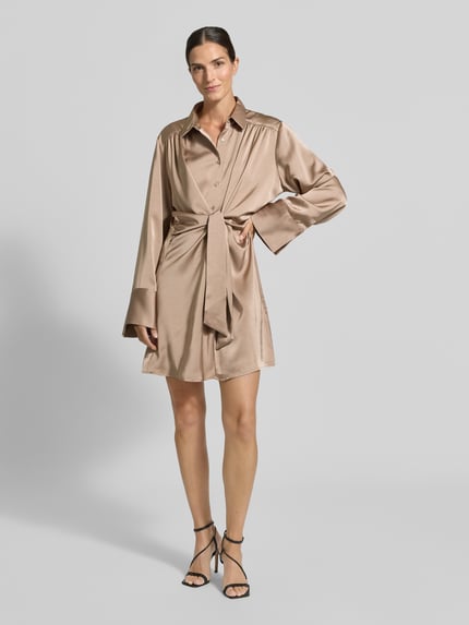 Frauenmodel trägt Knielanges Hemdblusenkleid mit Knotendetail Modell 'Biria' von Neo Noir aus glänzendem Satin in Beige, gerader Schnitt, Hemdblusenkragen, Ganzkörperansicht von vorne.