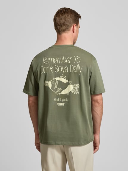 Männermodel trägt T-Shirt mit Label-Print Modell 'Baine' von WOODBIRD in Olivgrün, reine Bio-Baumwolle, Ganzkörperansicht Rückansicht.