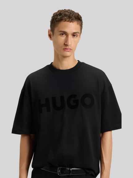 Męski model nosi Oversized T-Shirt aus reiner Baumwolle Modell 'DINKEE' von HUGO, jednolity czarny, krój oversize, nadruk z logo, dekolt okrągły, z krótkim rękawem, wykonany z bawełny, całkowity widok z przodu.