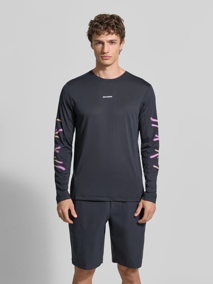 Męski model nosi Funktionsshirt mit Logo model 'Kout' od Salomon, czarny, krój regular fit, bluzka z długim rękawem, okrągły dekolt, długość tyłu w rozmiarze M: 70 cm, widok całej sylwetki z przodu.
