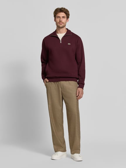 Männermodel trägt Regular Fit Troyer aus reiner Baumwolle von Lacoste in Dunkelrot, kombiniert mit gemusterten Hosen, Ganzkörperansicht von vorne. Herren Troyer von Lacoste