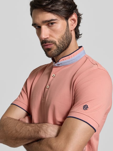 Męski model nosi Regular Fit Poloshirt aus reiner Baumwolle mit Stehkragen von Christian Berg Men w kolorze łososiowym z niebieską stójką i granatowymi detalami, krój regular fit, bawełna, krótkie rękawy, elastyczne zakończenia rękawów, długość tyłu 75 cm w rozmiarze M, Nahaufnahme górnej części ciała.