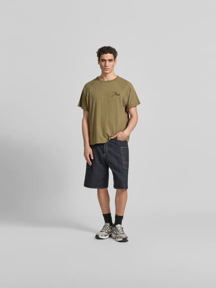 Męski model nosi Regular Fit T-Shirt aus reiner Baumwolle von RHUDE w kolorze oliwkowym, z wyhaftowanym logo, raglanowe rękawy, okrągły dekolt, długość tyłu 69 cm w rozmiarze M, fason krój regular fit, widok całej sylwetki z przodu.