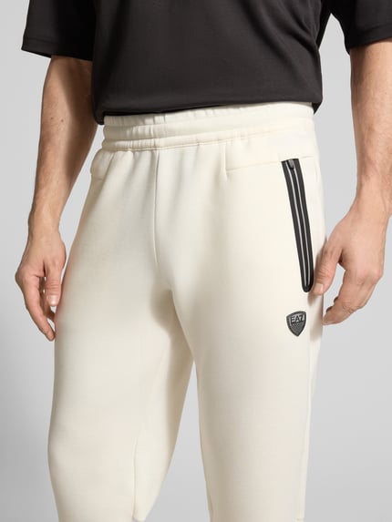 Männermodel trägt cremefarbene Sweatpants mit Viskose-Anteil Modell 'TRAIN PREMIUM SHIELD' von EA7 Emporio Armani, Nahaufnahme Oberkörper. Elastischer Bund, Reißverschlusstaschen und gerippte Abschlüsse, gerade Passform, detailreiche Materialstruktur.