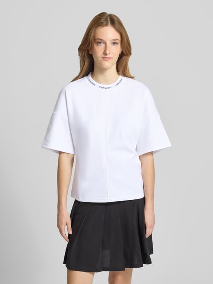 Frauenmodel trägt weißes Comfort Fit T-Shirt mit Label-Schriftzug von Calvin Klein Jeans, kombiniert mit schwarzem Rock, Ganzkörperansicht von vorne.