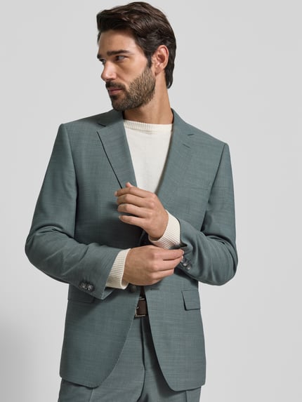 Męski model nosi Slim Fit Anzug aus Schurwoll-Mix Modell 'H-HUGE-2Pcs-253' od BOSS w jednolitej zielonej tonacji, tekstura gładka, komplet dwuczęściowy; widok całej sylwetki z przodu.