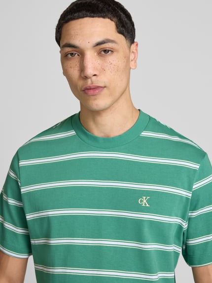 Męski model nosi t-shirt z wyhaftowanym logo od Calvin Klein Jeans, zielone paski, bawełna, prosty krój, okrągły dekolt, długość tyłu 69 cm, rozmiar M, zbliżenie na górną część ciała