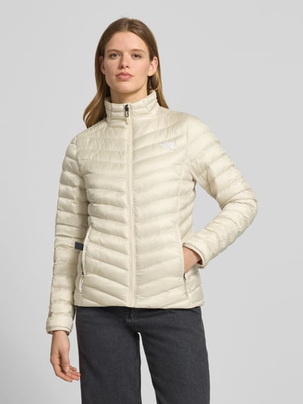 Frauenmodel trägt Steppjacke mit Stehkragen und Label-Stitching von The North Face in Creme, Reißverschlusstaschen, glatte Struktur, Ganzkörperansicht von vorne, Regular Fit, Rückenlänge 62 cm, Schulterbreite 38 cm, Brustweite 46 cm, Ärmellänge 64 cm.