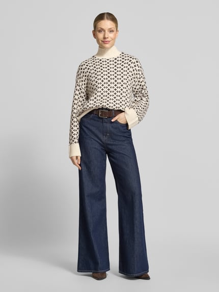 Frauenmodel trägt Pullover mit Jacquardstruktur Modell 'VMBIRCH' von VERO MODA, cremefarbener Strickstoff mit grafischem Allover-Muster, dunkle Jeans, ganzheitliches Muster und gerippte Abschlüsse, Ganzkörperansicht von vorne.