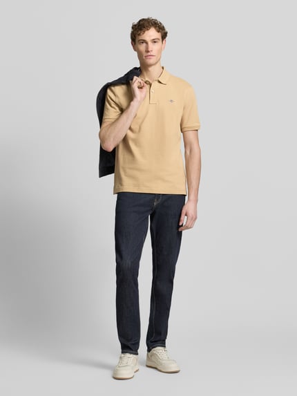 Männermodel trägt Slim Fit Poloshirt mit Logo-Stitching von Gant in Beige, reine Baumwolle, Polo-Kragen, dunkle Jeans, Ganzkörperansicht von vorne.