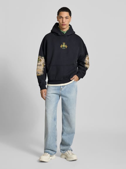 Männermodel trägt Oversized Hoodie aus reiner Baumwolle van RHUDE in zwart met labelprints, kangoeroezak, ribboorden, ruglengte bij maat M 63 cm, Ganzkörperansicht von vorne.