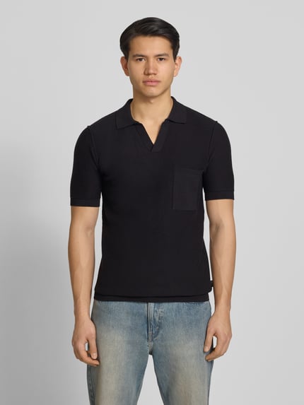 Männermodel trägt Slim Fit Poloshirt in Strick-Optik von Replay in Schwarz mit V-Ausschnitt und Brusttasche, Ganzkörperansicht von vorne.