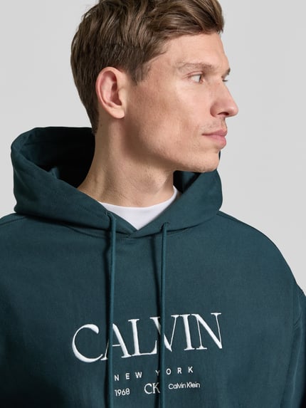 Männermodel trägt Hoodie mit Label-Print und Känguru-Tasche von CK Calvin Klein in Dunkelgrün, Baumwoll-Elasthan-Mix, Nahaufnahme Oberkörper.