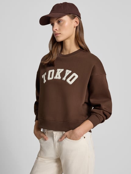 Frauenmodel trägt Relaxed Fit Sweatshirt aus Baumwoll-Mix Modell 'Tatum' von JJXX in Braun mit Statement-Patch, Rippbündchen, überschnittenen Schultern und Rundhalsausschnitt, Ganzkörperansicht von vorne.
