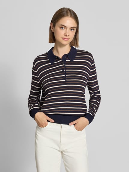 Modelka nosi koszulkę polo 'Poloshirt mit geripptem Saum' od REVIEW, mieszanka materiałów z bawełną, prosty krój, długi rękaw, kołnierzyk polo, prążkowany dół, długość tyłu w rozmiarze S: 57 cm, długość rękawa w rozmiarze S: 62 cm, paski granatowe, kremowe i beżowe, widoczne faktury prążkowania, widok całej sylwetki z przodu.