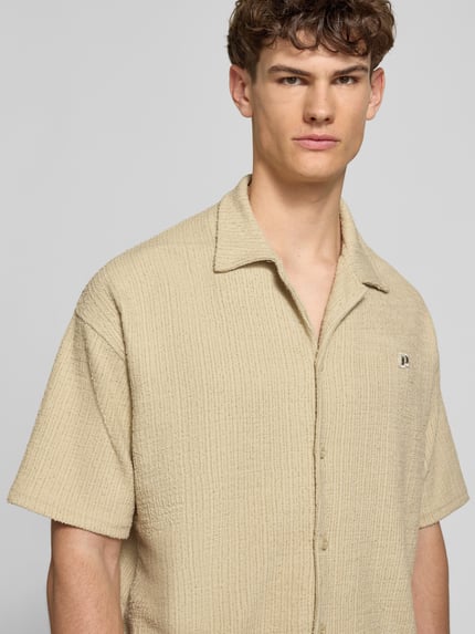 Männermodel trägt Regular Fit Freizeithemd mit Label-Patch Modell 'LIBCO' von Pegador, beigefarbener Material-Mix, Knopfleiste, Reverskragen, Kurzärmelig, Struktur-Oberfläche, Nahaufnahme Oberkörper. Rückenlänge bei Größe M: 72 cm, Schulterbreite: 41 cm, Ärmellänge: 35 cm.