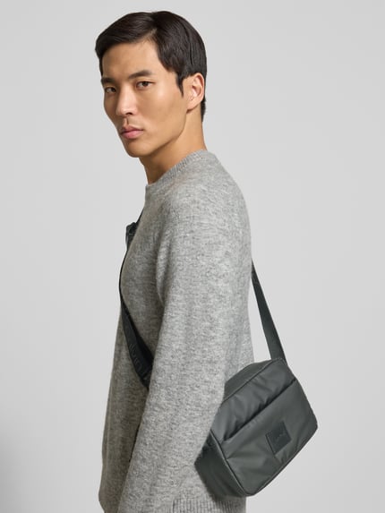 Mannelijk model draagt Handtasche mit Label-Patch und verstellbarem Schulterriemen van CK Calvin Klein, grijs, utility-look, Ganzkörperansicht van voren. Hoofdvak met ritssluiting, labelpatch, verstelbare schouderband, breedte 30 cm, lengte 16 cm, diepte 7 cm, bandlengte 145 cm.