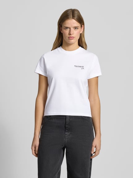Kobiece modelka nosi damski t-shirt z bawełny o prostym kroju, okrągłym dekolcie i detalu z logo Carhartt Work In Progress, długość tyłu 49 cm, rozmiar S, widok całej sylwetki z przodu.