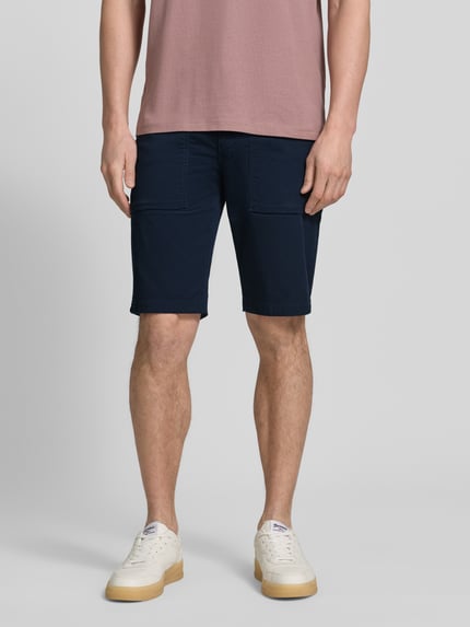Męski model nosi Regular Fit Cargoshorts aus Baumwoll-Mix von s.Oliver BLACK LABEL, granatowy twill, 5 kieszeni, szerokie szlufki, naszywka z logo, zapięcie na guzik i ukryty zamek, widok całej sylwetki z przodu.