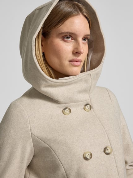 Vrouwenmodel draagt Wollmantel mit Kapuze van VERO MODA, effen lichtbeige, dubbele knoopsluiting, capuchon, zijzakken, lange mouwen, afgeronde zoom, close-up bovenlichaam.