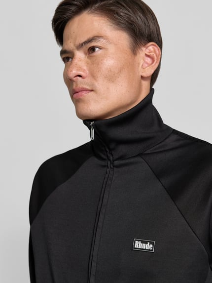 Männermodel trägt Sweatjacke mit Stretch-Anteil von RHUDE, Schwarz, Label-Print, Zweiwege-Reißverschluss, Raglanärmel, Nahaufnahme Oberkörper.