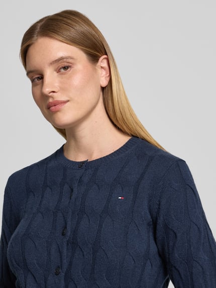 Frauenmodel trägt Strickjacke aus Wolle mit Logo-Detail von Tommy Hilfiger in Dunkelblau mit Zopfmuster, Nahaufnahme Oberkörper.