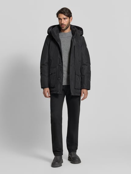 Męski model nosi Kurtka 'Jacke mit Sorona® Aura Filling' od NAUTGÅRD, czarny kolor, krój regular fit, kaptur, stójka, oddychający materiał, wodoodporny, wypełnienie Sorona®, kieszenie z patką, detal z logo, zapięcie na zatrzask, długość tyłu w rozmiarze M: 82 cm, szerokość ramion w rozmiarze M: 47 cm, szerokość w rozmiarze M: 51 cm, długość rękawów w rozmiarze M: 69 cm, zdjęcie całej sylwetki z przodu.