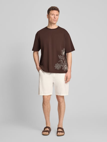 Mannelijk model draagt Loose Fit T-shirt met Stitchings van Scotch & Soda, bruin met witte print, puur katoen, ronde hals, ruglengte maat M 71 cm, loose fit, ganzekörperaanzicht van voren.