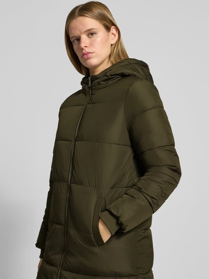 Frauenmodel trägt Puffermantel mit Kapuze von PIECES in Olivgrün, Longline-Schnitt, mit Reißverschluss, Teilungsnähten und Taschen, Nahaufnahme Oberkörper.