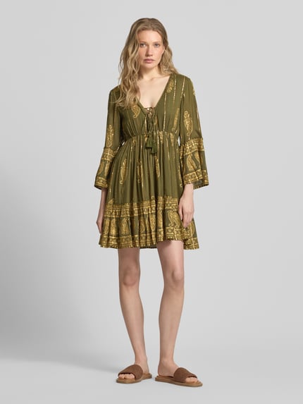 Frauenmodel trägt Minikleid aus Viskose-Mix mit Schnürung von Apricot in olivgrün mit goldfarbenem Muster, A-Linie, V-Ausschnitt, Trompetenärmel, Ganzkörperansicht von vorne, Rückenlänge 83 cm, Brustweite 35 cm, Ärmellänge 50 cm.