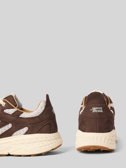 Produktfoto, Sneaker mit Kontrastbesatz von The Mercer, braune und cremefarbene Oberfläche, strukturierte Sohle, Label-Print, abgerundete Form, Schnürverschluss, Größenangaben zu Absatzhöhe, Schafthöhe und Schaftweite, neutrale Hintergrundfarbe.