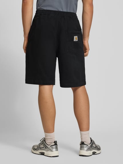Männermodel trägt Shorts aus Baumwoll-Mix Modell 'Floyde' von Carhartt Work In Progress in Schwarz, lockerer Schnitt, Ganzkörperansicht Rückansicht. Kontext: Herren Shorts mit elastischem Bund, Tunnelzug, Eingrifftaschen, Bundweite M 39 cm, Innenbeinlänge M 25 cm.