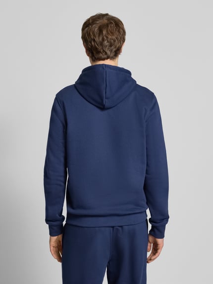 Männermodel trägt Hoodie mit Kapuze von adidas Originals aus Katoenmix, los model, capuchon, tunnelkoord, labelstitching, kangoeroezak, ribboorden, ruglengte maat M 68 cm, mouwlengte maat M 69 cm, effen donkerblauw, zachte textuur. Ganzkörperansicht Rückansicht.