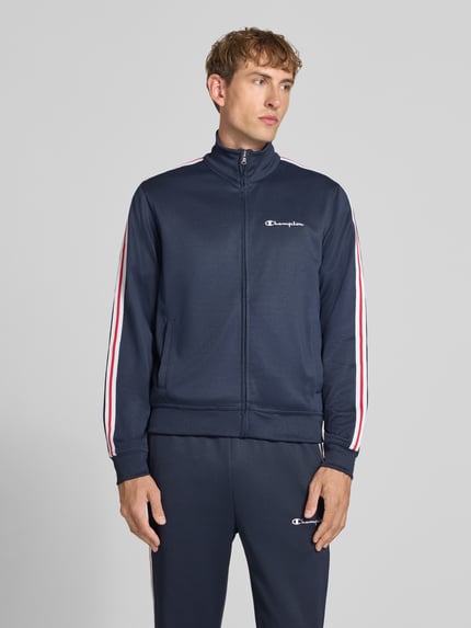 Männermodel trägt Sweatjacke mit Bordüre von Champion im Baumwoll-Mix, gerader Schnitt, dunkelblau mit roten und weißen Streifen, Nahaufnahme Oberkörper.