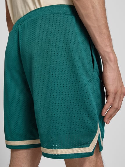 Mężczyznamodel nosi szorty mit Label-Stitching und elastischem Bund von PROHIBITED, siateczka, krój relaxed fit, szerokość w rozmiarze M: 41 cm, długość wewnętrzna nogawki w rozmiarze M: 18 cm, zielony kolor, kremowe detale, tekstura siateczki, detailaufnahme tylnej części.