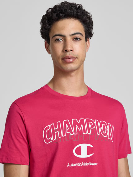 Männermodel trägt T-Shirt mit Label-Print und Rundhalsausschnitt von CHAMPION in Rot, reine Baumwolle, Regular Fit, Nahaufnahme Oberkörper.
