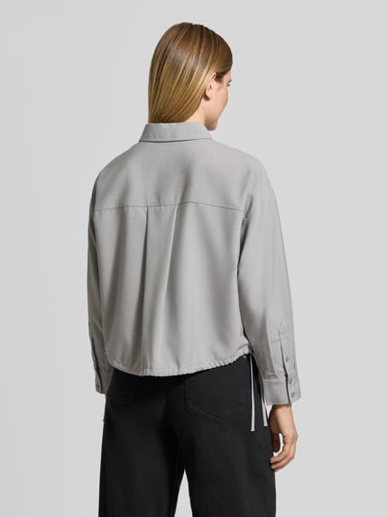 Frauenmodel trägt Cropped Langarmbluse mit Tunnelzugsaum Modell 'Felanga' von OPUS in grauer Melange-Optik und Strukturmuster, Webware mit Knöpfen, Brusttasche, überschnittenen Schultern, langen Ärmeln mit Manschetten, Tunnelzug am Saum, Produktfoto in Ganzkörperansicht Rückansicht.
