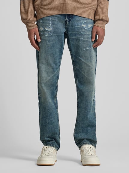 Männermodel trägt Tapered-Fit Jeans aus Baumwoll-Mix Modell 'TROY' von BOSS Orange in blauer Denim-Optik mit Destroyed-Details, Label-Patch, Nieten, 5-Pocket-Design, Knopf und verdecktem Reißverschluss, Ganzkörperansicht von vorne.