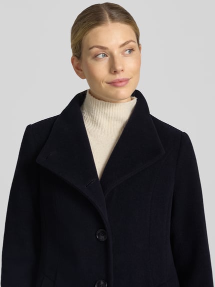 Vrouwenmodel draagt Regular fit lange jas met opstaande kraag model 'NADA' van Milo Coats, materiaalmix met scheerwol, knoopsluiting, steekzakken, ruglengte maat 36: 118 cm, schouderbreedte 40 cm, borstwijdte 47 cm, mouwlengte 67 cm, donkere kleur, zachte textuur, Nahaufnahme Oberkörper.