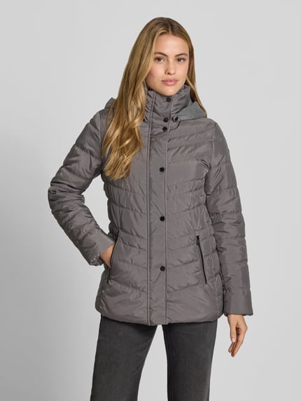 Modelka nosi 'Jacke in Stepp-Optik mit Kapuze' od FUCHS SCHMITT w kolorze szarym, z efektem pikowania, taliowany krój, kaptur, ocieplana stójka, wpuszczane kieszenie na zamek błyskawiczny, długość tyłu w rozmiarze 36: 74 cm, szerokość ramion w rozmiarze 36: 40 cm, szerokość klatki piersiowej w rozmiarze 36: 48 cm, długość rękawów w rozmiarze 36: 64 cm, ujęcie całej sylwetki od przodu.